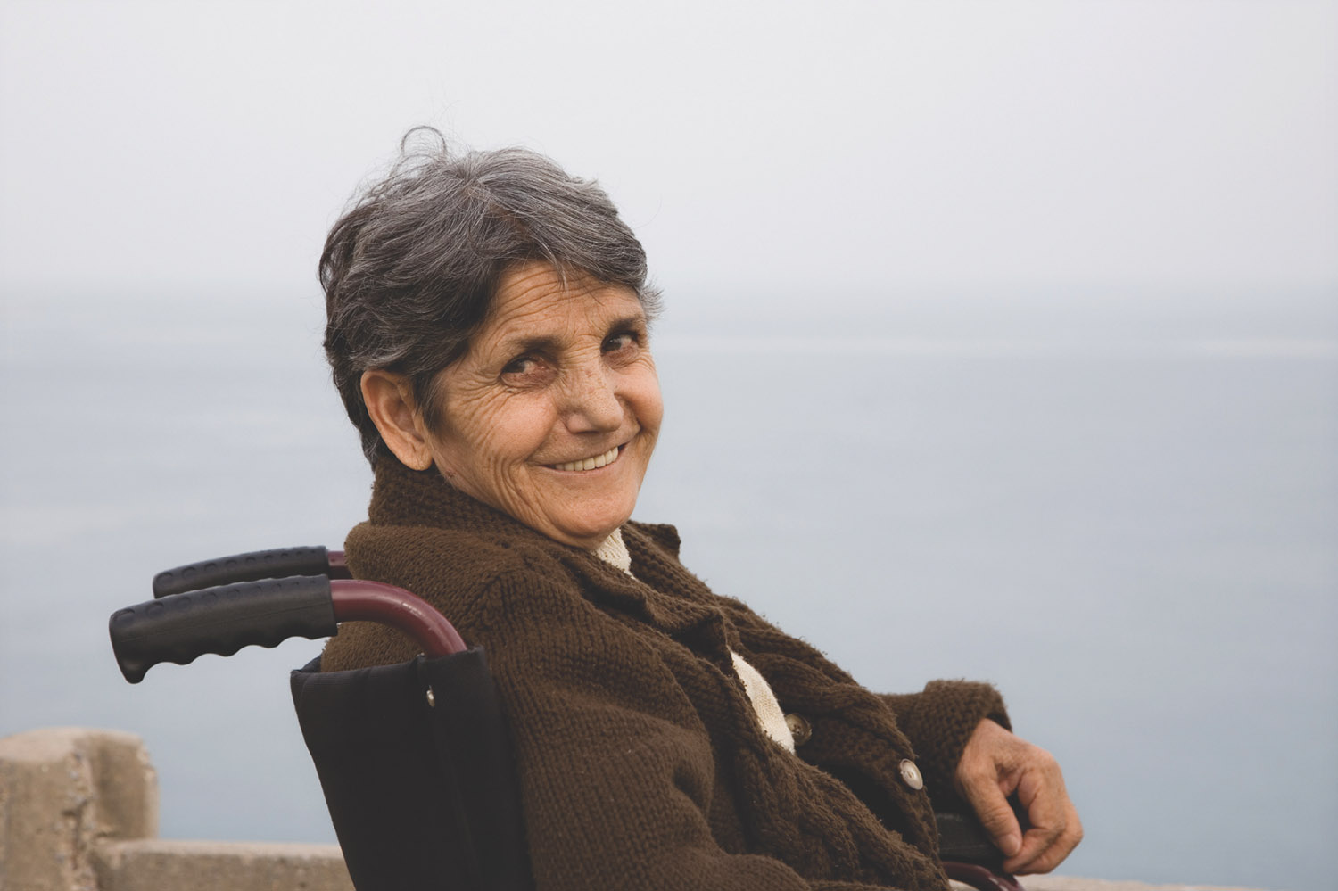A senior woman in a wheelchair smiling at camera with a beach in the background