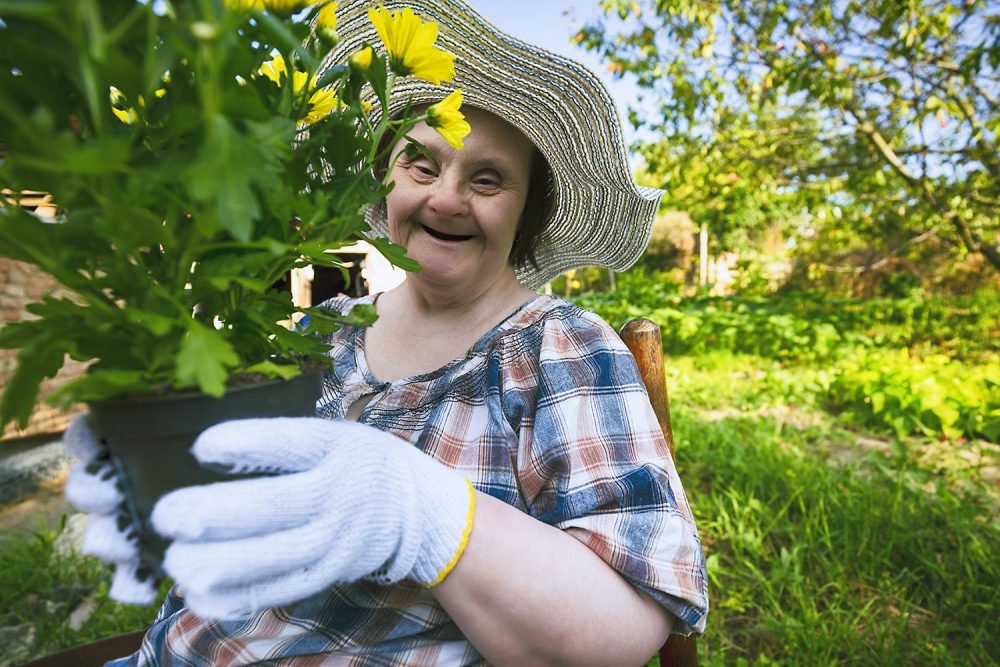 A woman living with down syndrome is working in the garden and holding a pot with flowers